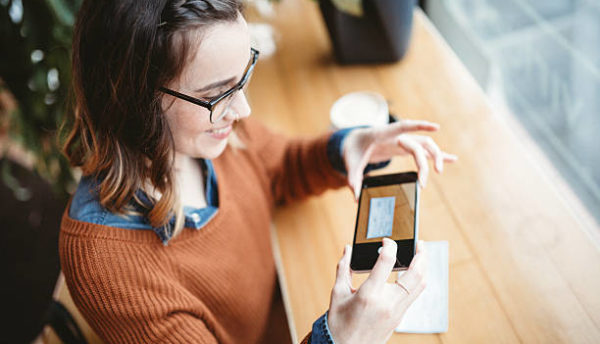 A smiling young woman takes a picture with her smart phone of a check or paycheck for digital electronic depositing, also known as