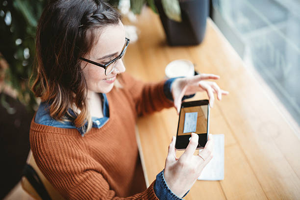 A smiling young woman takes a picture with her smart phone of a check or paycheck for digital electronic depositing, also known as "Remote Deposit Capture". She sits in a coffee shop, enjoying an espresso latte. Bright sunlight shines in the window.